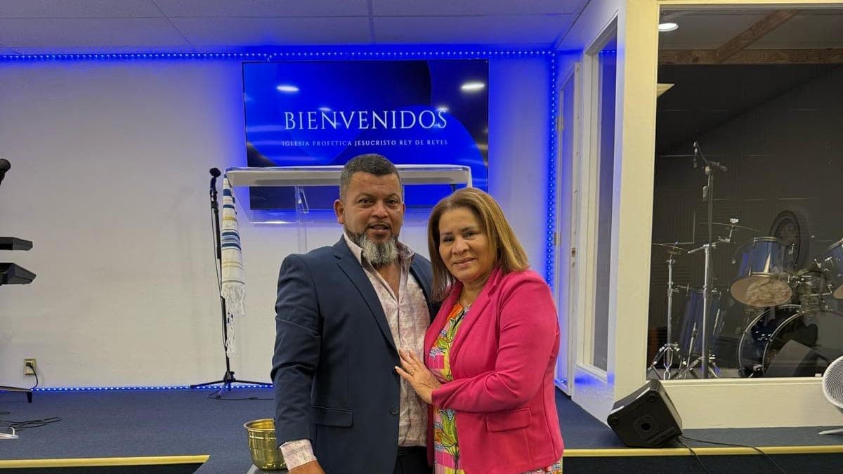 Los pastores Elmer y Carla Reyes de pie frente al altar, con la pantalla de bienvenida de la iglesia detrás